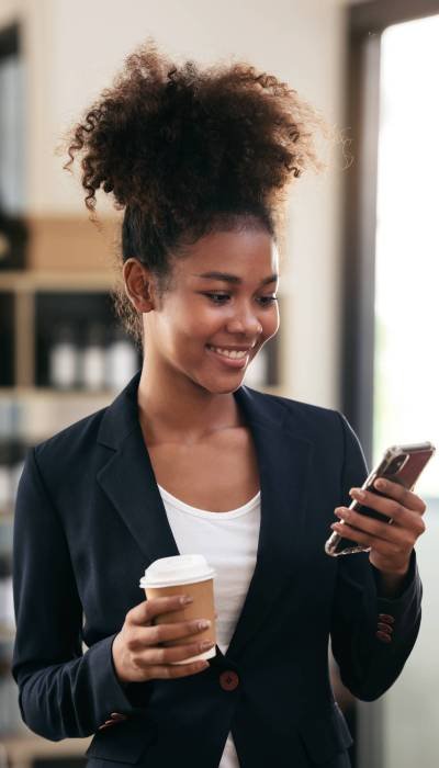 African american businesswoman in suit drinking coffee and reading accounting information of new business on smartphone while standing to working and thinking strategy of project in modern workspace.